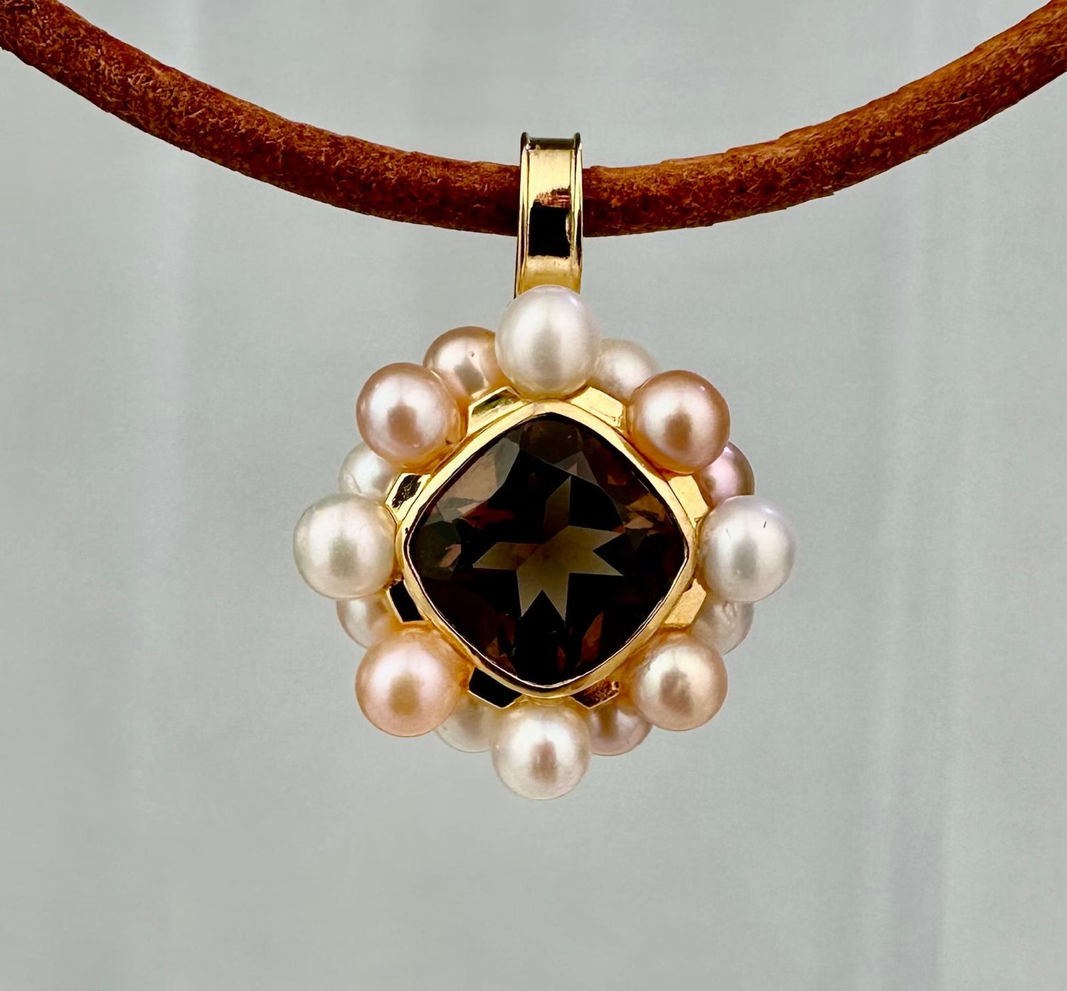 Gold pendant with central smokey quartz stone and freshwater pearl on a brown leather cord against a light gray background